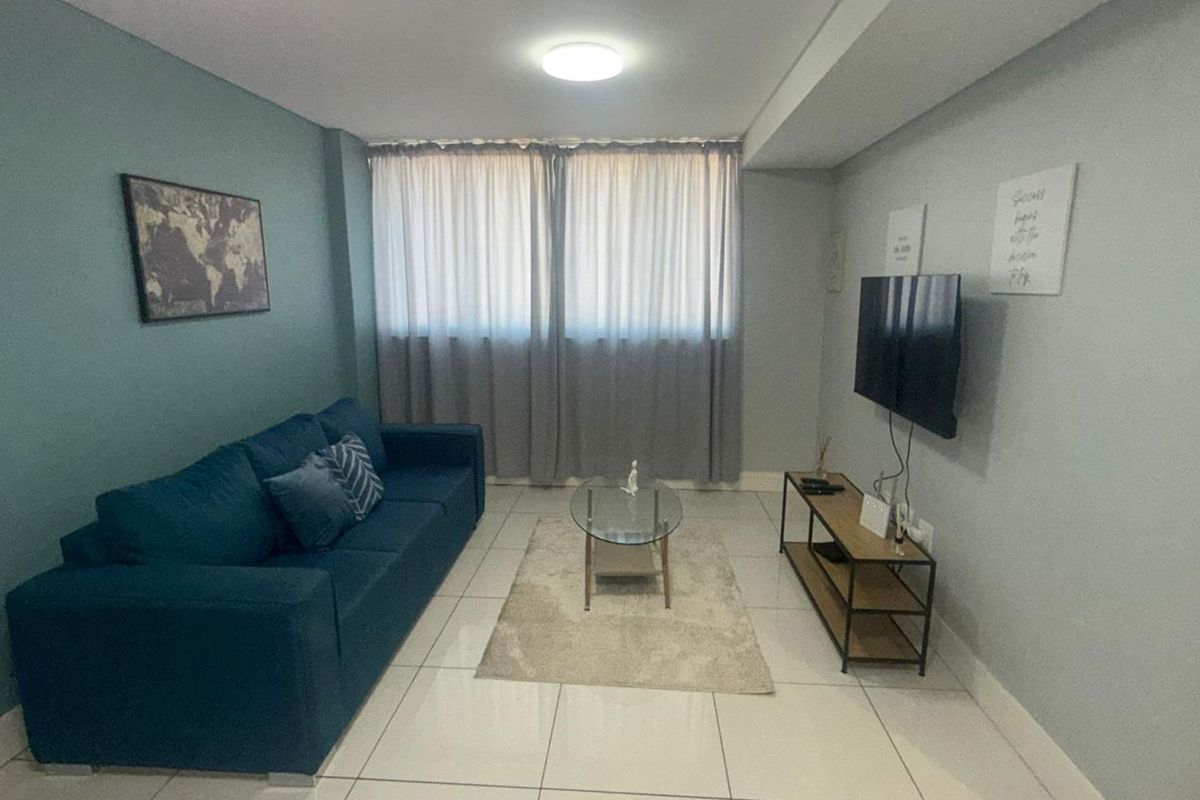 A well-maintained living room featuring neutral tiled flooring, a soft teal accent wall, and large windows with curtains for natural light.