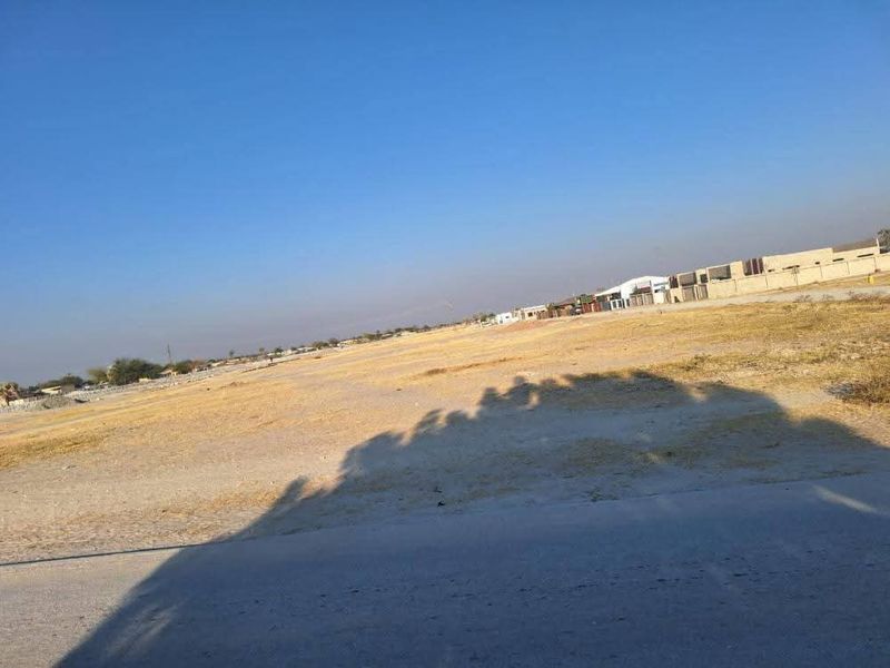 A wide-angle view of a vacant residential plot of land with dry vegetation and surrounding neighborhood development in the distance.