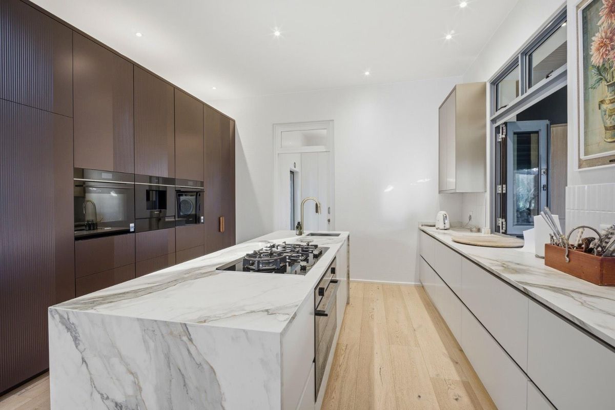 Modern kitchen featuring a large marble-topped island with integrated gas cooktop, sleek dark cabinetry, built-in ovens, and light timber flooring.
