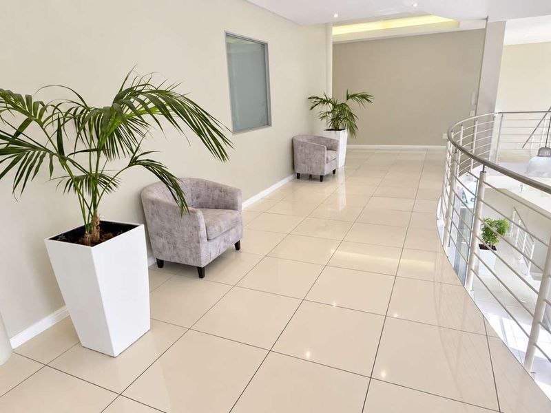 A bright and spacious office hallway featuring polished tile flooring, recessed lighting, and a modern stainless steel balustrade overlooking the lower level.