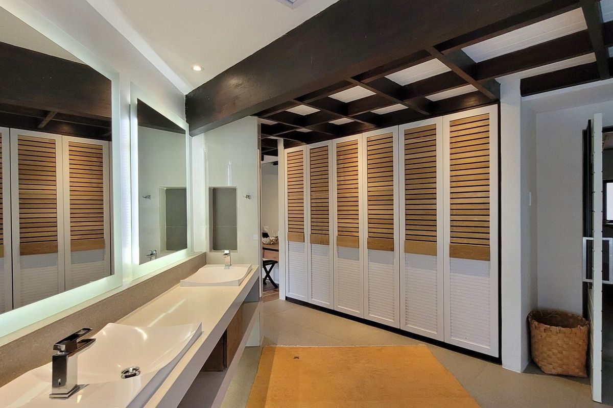 Modern bathroom featuring a double vanity with vessel sinks, backlit mirrors, and extensive built-in louvered cabinetry under a decorative exposed timber ceiling.