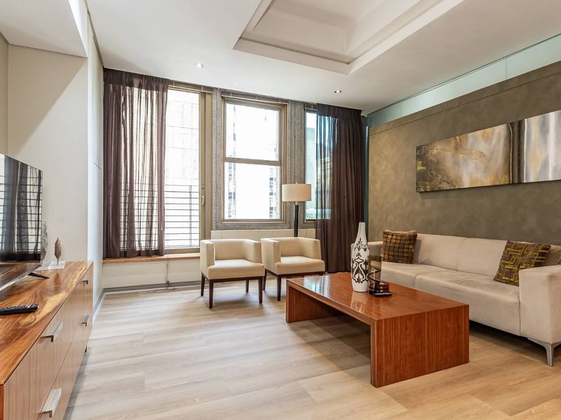 A well-maintained living room featuring modern laminate flooring, a recessed ceiling, and a textured accent wall with large windows providing natural light.
