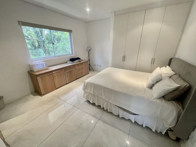 A bright bedroom featuring large-format tiled flooring, extensive built-in cupboards, and a window overlooking greenery.