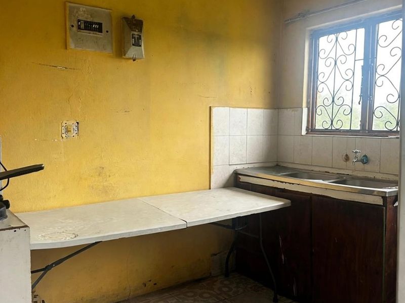 A basic kitchen area featuring tiled flooring, a stainless steel sink, and a security-grilled window, requiring significant cosmetic renovation and surface repairs.