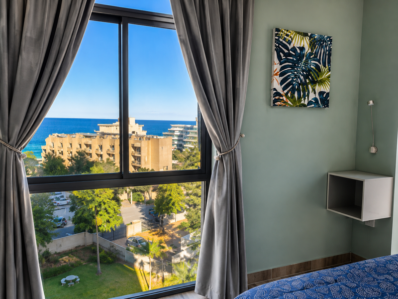 A bright bedroom featuring a large sliding window with expansive coastal and neighbourhood views, complemented by a wall-mounted light and built-in shelving.