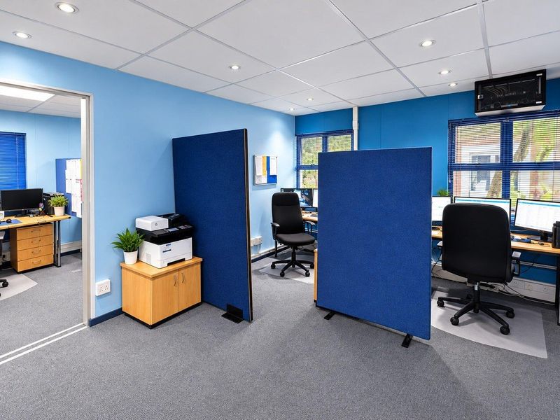 A functional office space featuring a suspended ceiling with recessed lighting, carpeted flooring, and multiple workstations separated by blue privacy partitions.