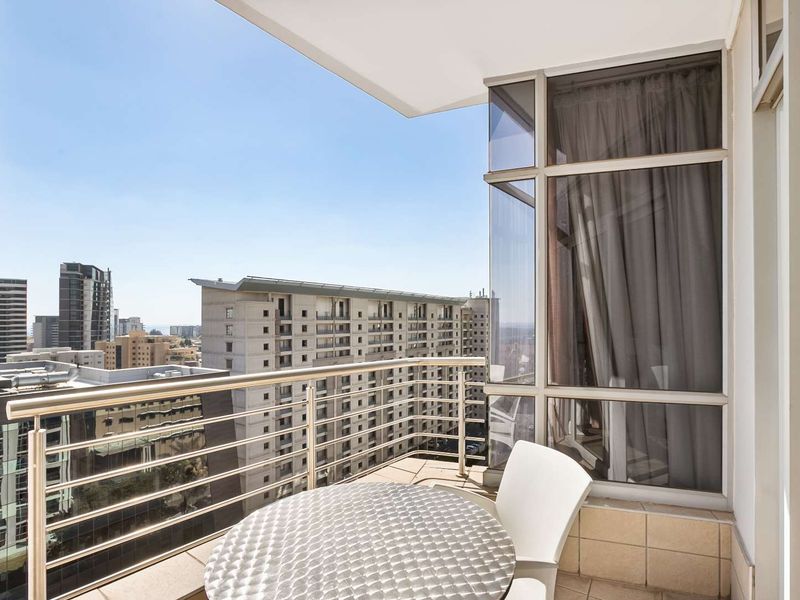 A modern apartment balcony featuring a stainless steel balustrade, tiled flooring, and expansive glass windows offering an elevated urban city view.
