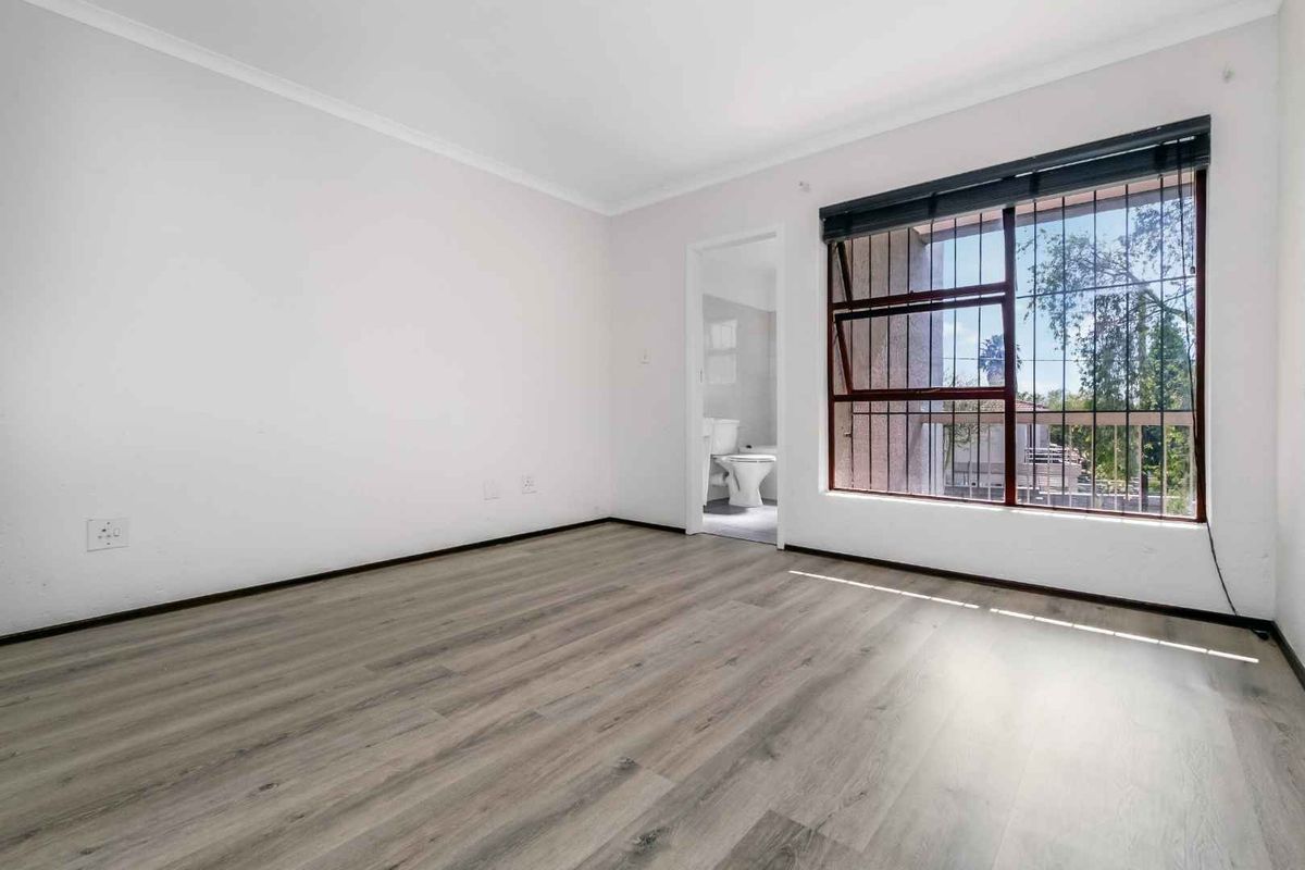 A bright, empty bedroom featuring modern wood-look laminate flooring, a large window with security bars, and direct access to an en-suite bathroom.