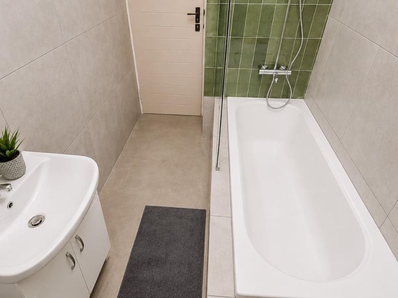 A modern bathroom featuring a white vanity with basin, a bathtub with a glass shower screen, and stylish green accent tiling.