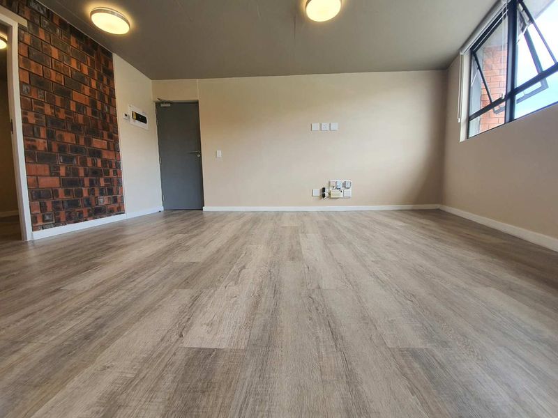 A bright, empty living room featuring modern laminate flooring, an exposed brick accent wall, and large windows providing natural light.