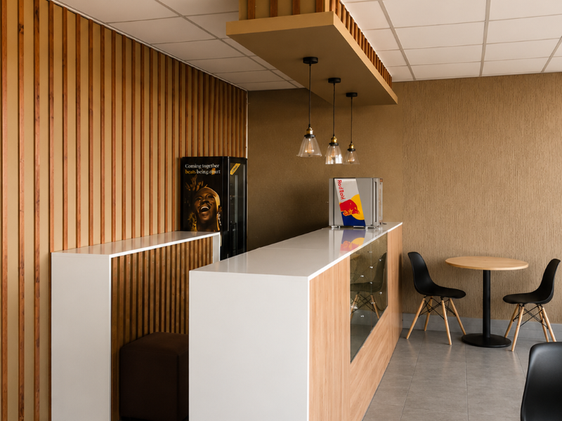 A modern retail bar area featuring a sleek white and wood-finish counter, timber slat wall accents, pendant lighting, and a clean tiled floor.