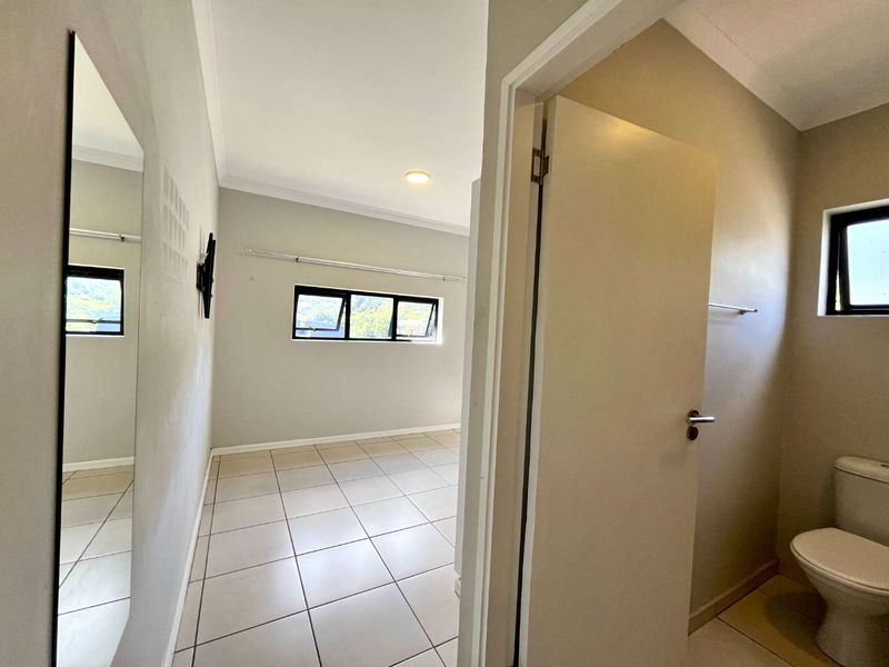 A bright, empty bedroom featuring neutral tiled flooring, aluminium window frames, and an en-suite bathroom area with a built-in toilet.