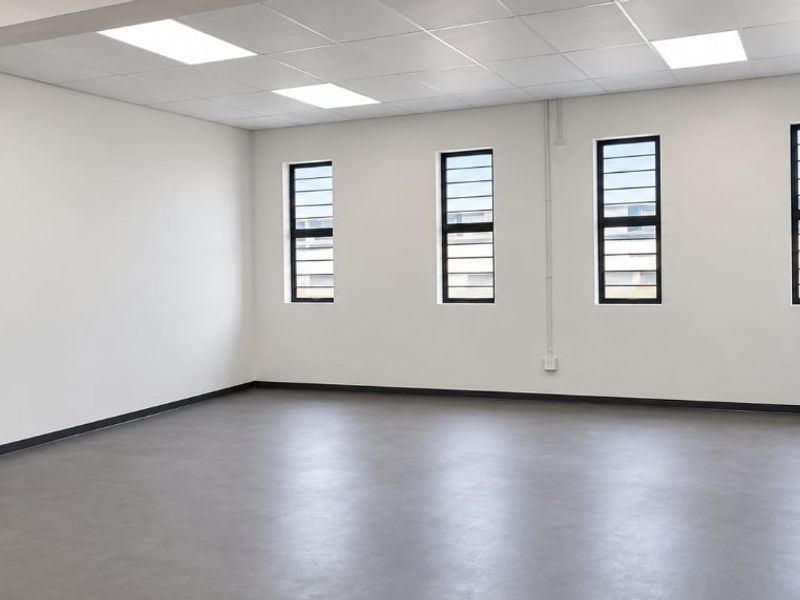 A bright, open-plan office space featuring neutral walls, durable vinyl flooring, suspended ceiling with recessed lighting, and large windows with security bars.