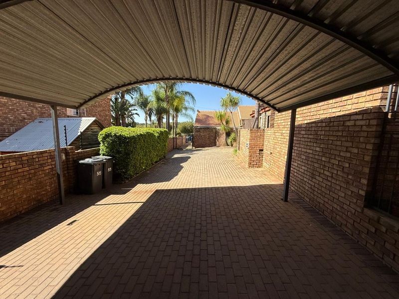 A paved driveway area featuring a covered carport structure with facebrick boundary walls and established garden greenery.