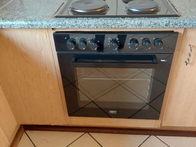 A functional kitchen area featuring a built-in oven, electric hob, granite countertops, and wooden cabinetry with tiled flooring.