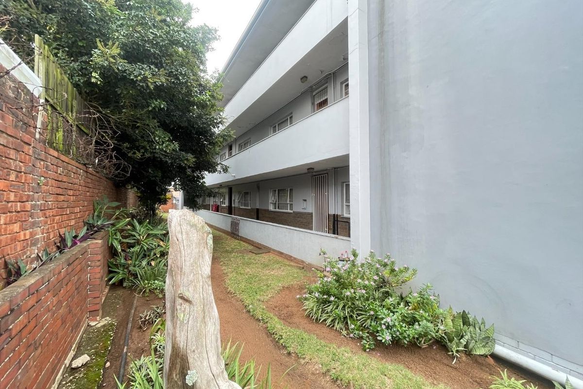 Rear exterior view of the apartment building featuring a concrete walkway, landscaped garden area, and brick boundary wall.