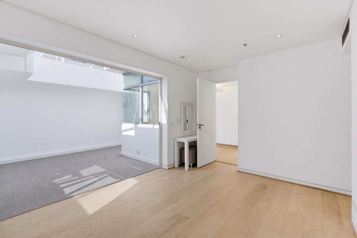 A bright, modern bedroom featuring hardwood floors, recessed lighting, and a large skylight, with an adjoining carpeted area accessible through a sliding glass door.