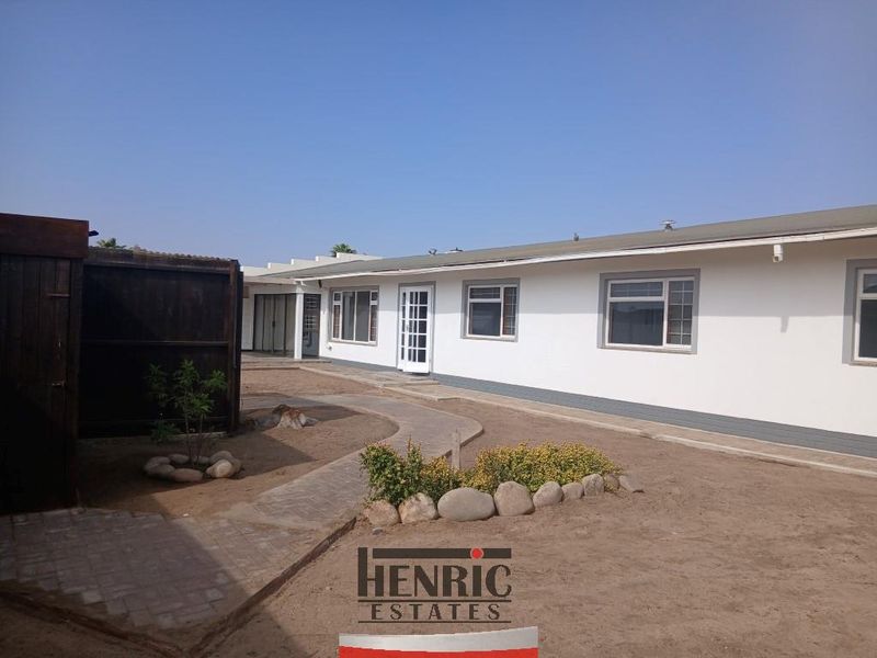 Rear view of the property featuring a paved walkway, white exterior walls, and aluminium window frames, set against an unlandscaped garden area.