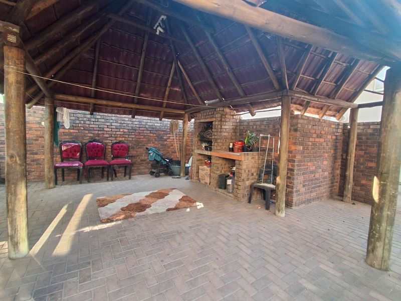 Covered outdoor patio area featuring a thatched roof, timber support poles, and a built-in brick braai, perfect for outdoor entertaining.
