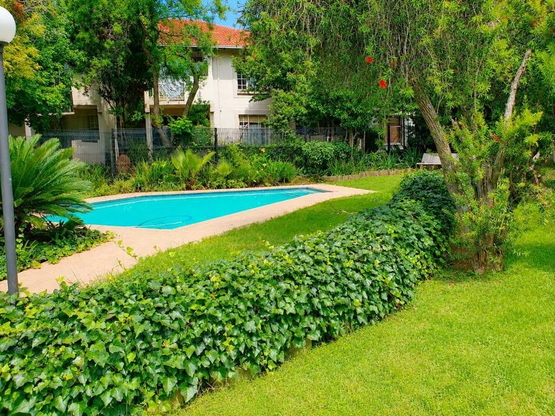 A well-maintained communal swimming pool area surrounded by lush green lawns, mature trees, and established garden landscaping.
