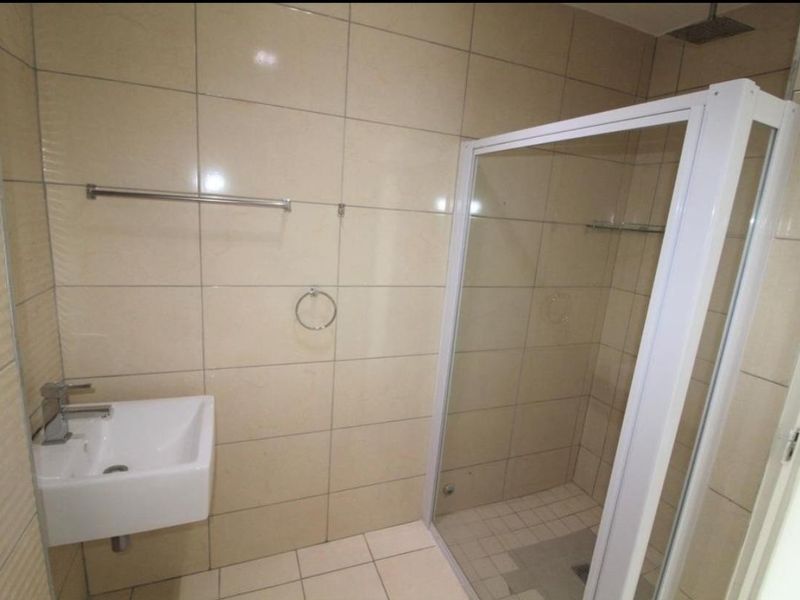 A clean, modern bathroom featuring a wall-mounted basin, a glass-enclosed shower with a rain shower head, and neutral floor-to-ceiling wall tiles.