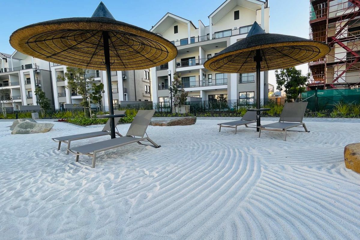 A well-maintained communal outdoor sand area with thatched umbrellas and landscaped surroundings within an apartment complex.
