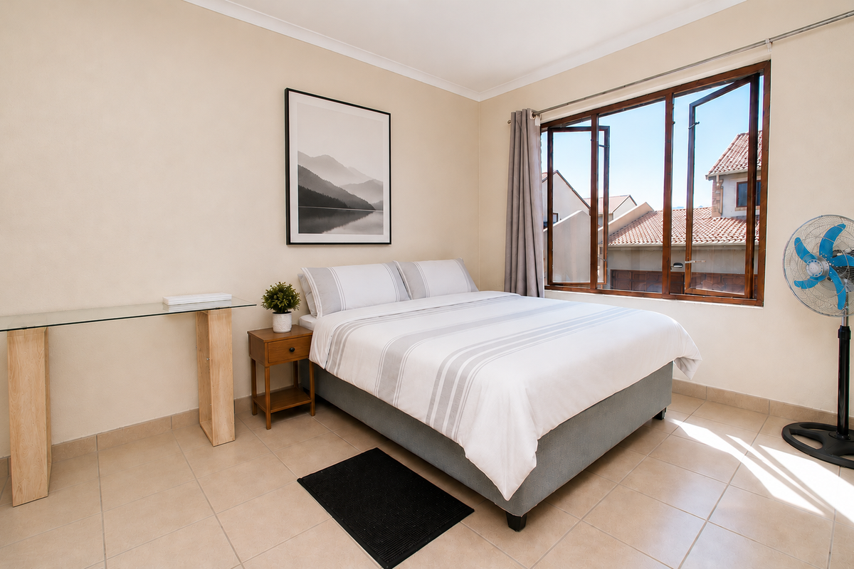 A bright bedroom featuring tiled flooring, neutral walls, and a large window providing natural light and views of the surrounding townhouse complex.