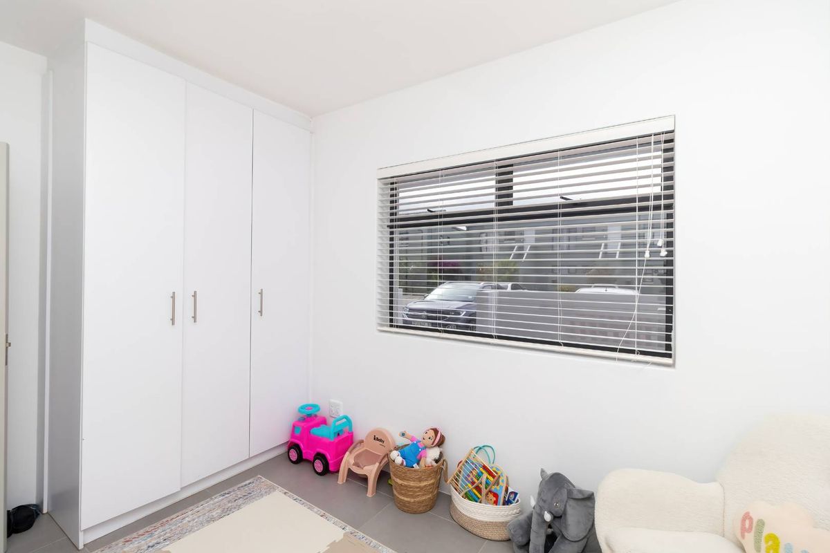 A bright bedroom featuring neutral tiled flooring, built-in white cupboards, and a window fitted with venetian blinds.