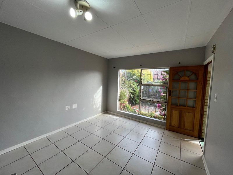 An empty living room featuring tiled flooring, a timber entrance door, and a large window with security bars overlooking the garden.