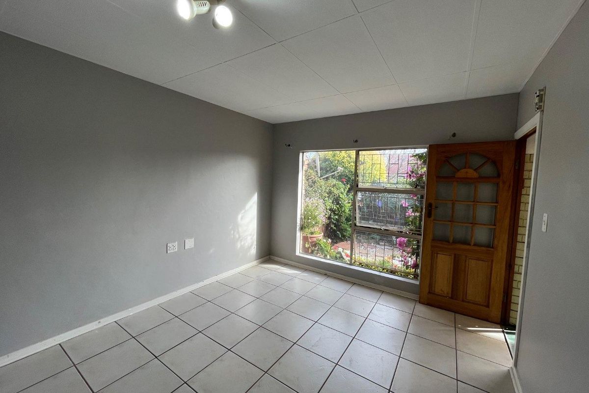 An empty living room featuring tiled flooring, a timber entrance door, and a large window with security bars overlooking the garden.