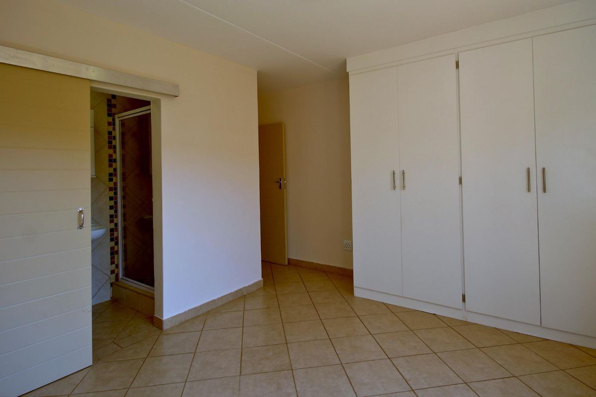 A spacious, empty bedroom featuring neutral tiled flooring, ample built-in cupboard storage, and direct access to an en-suite bathroom via a sliding door.