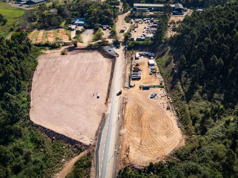 Aerial view of a large industrial development site featuring cleared land, a paved access road, and surrounding natural forest landscape.