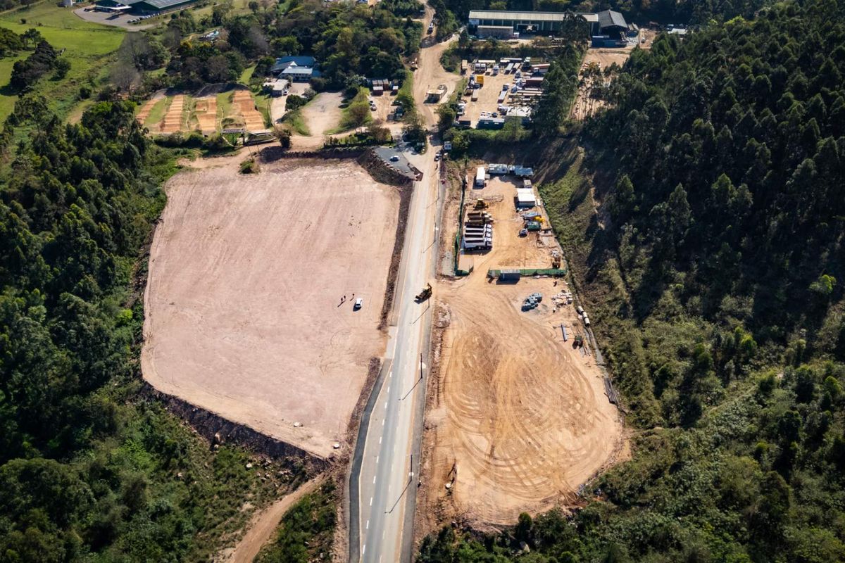 Aerial view of a large industrial development site featuring cleared land, a paved access road, and surrounding natural forest landscape.