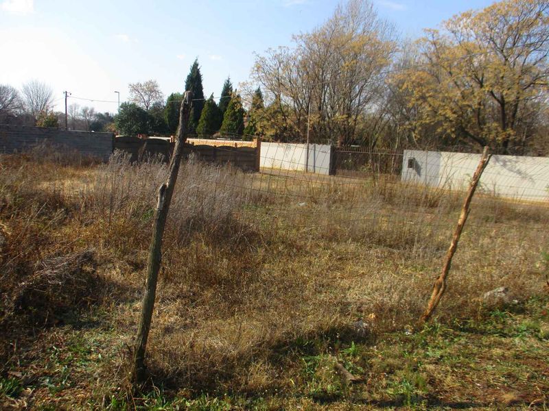 A vacant residential plot featuring overgrown grass, perimeter walling, and basic wire fencing with a metal gate.