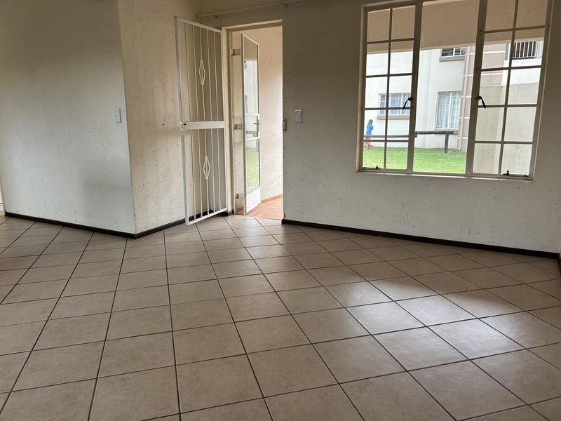 An empty living area featuring ceramic floor tiles, a security gate, and large steel-framed windows looking out onto the complex grounds.