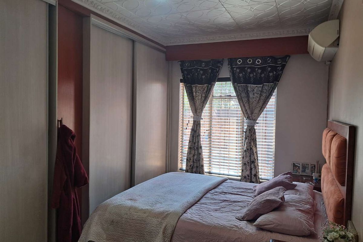 A bedroom featuring built-in sliding door cupboards, a wall-mounted air conditioning unit, and a window with fitted blinds.