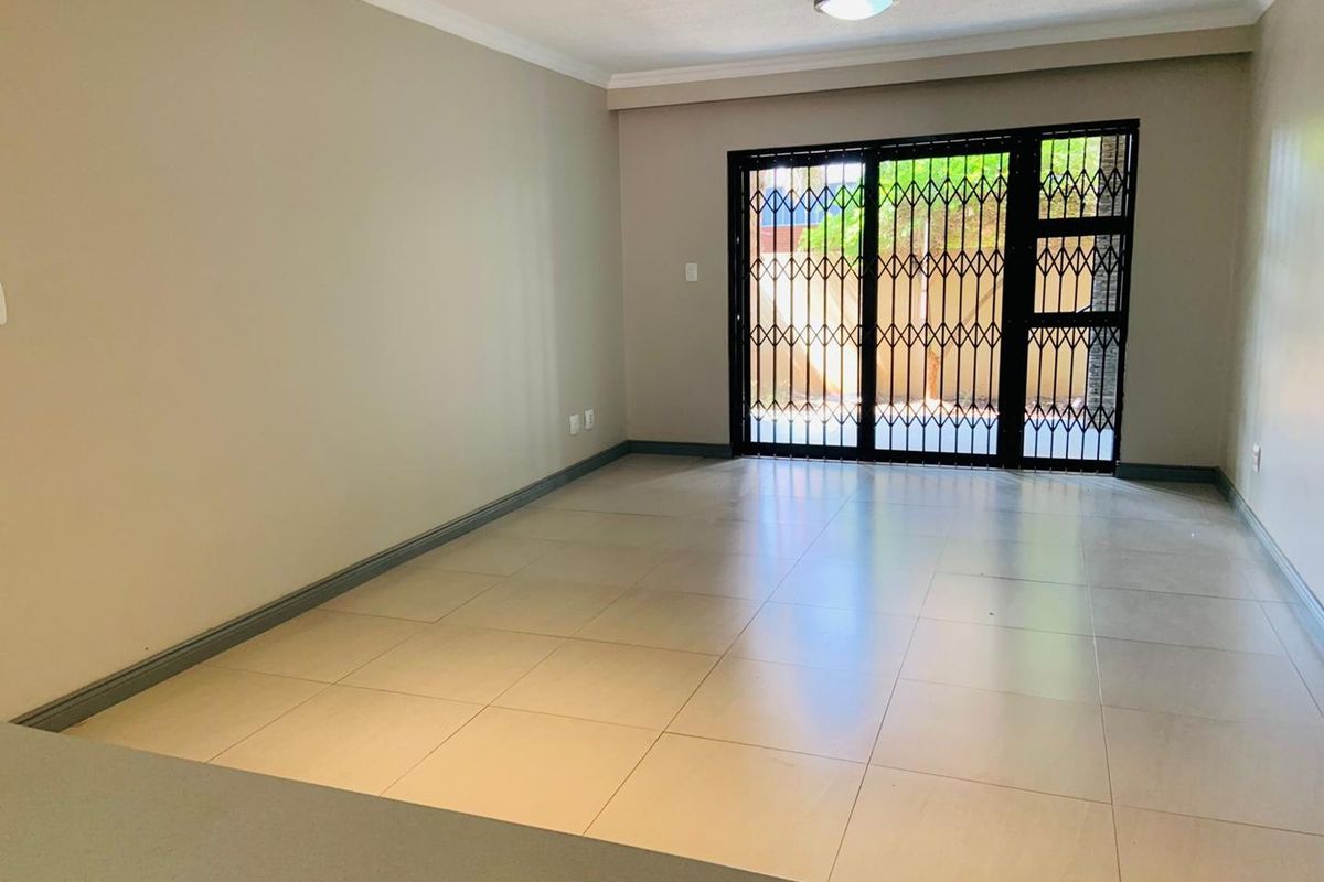 A spacious, empty living area featuring neutral tiled flooring, coved ceilings, and a sliding glass door secured with a black metal security gate.