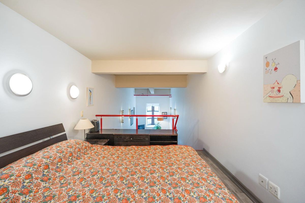 A mezzanine bedroom area featuring laminate flooring, wall-mounted lighting, and a red metal safety balustrade overlooking the lower level.