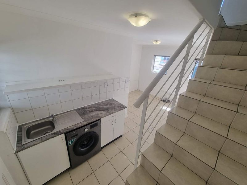 A compact kitchen area featuring a stainless steel sink, tiled splashback, and built-in cabinetry, adjacent to a tiled staircase with a metal balustrade.