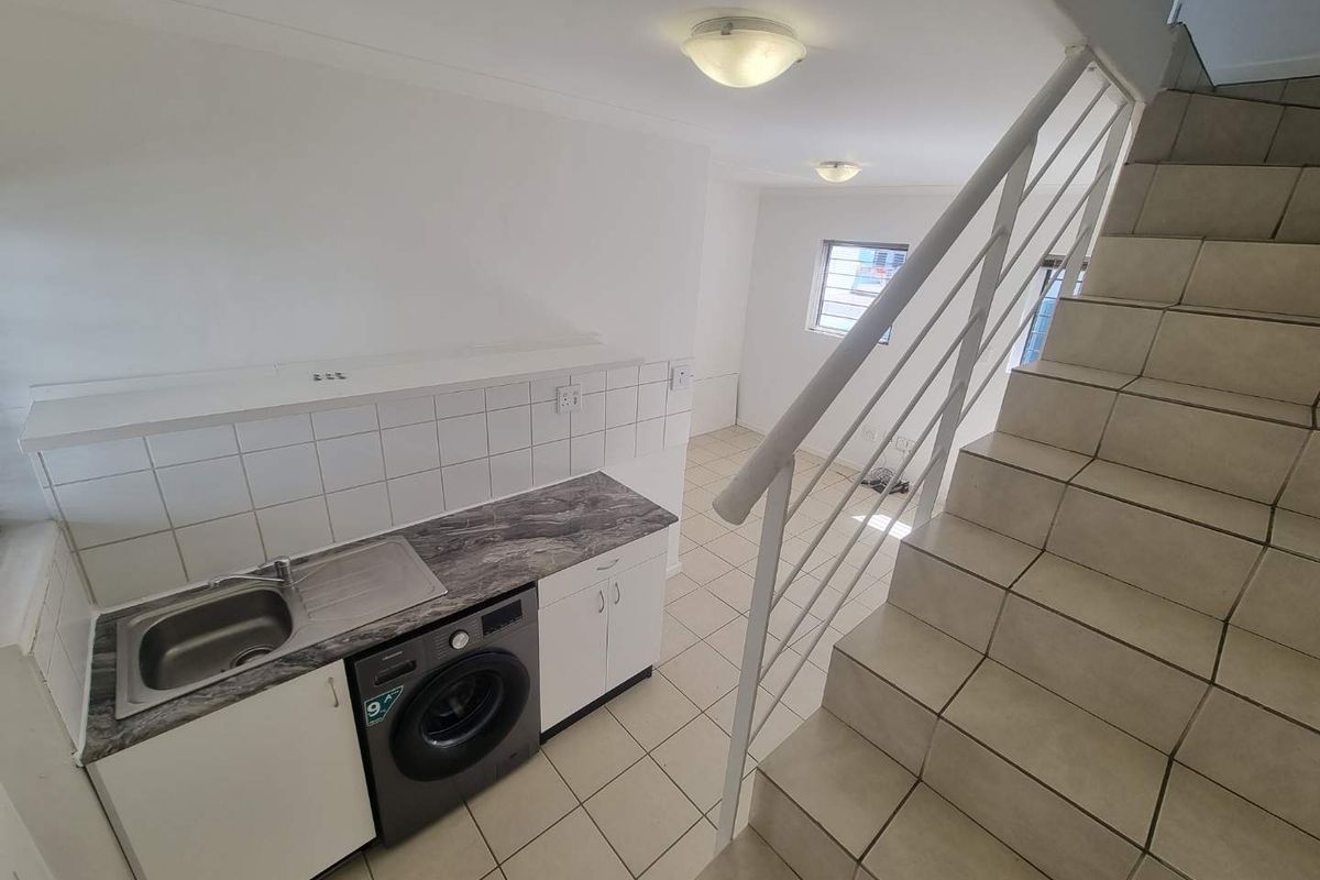 A compact kitchen area featuring a stainless steel sink, tiled splashback, and built-in cabinetry, adjacent to a tiled staircase with a metal balustrade.
