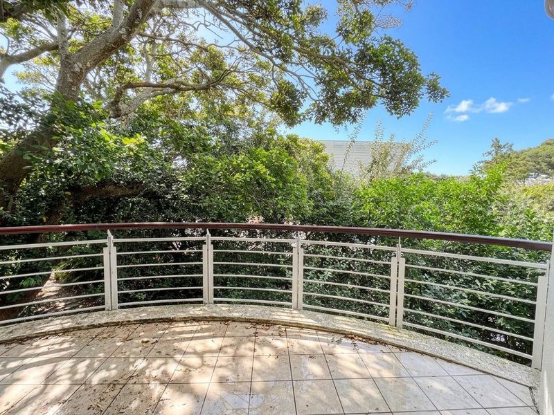 A curved balcony featuring tiled flooring and a metal balustrade with a wooden handrail, overlooking mature trees and greenery.