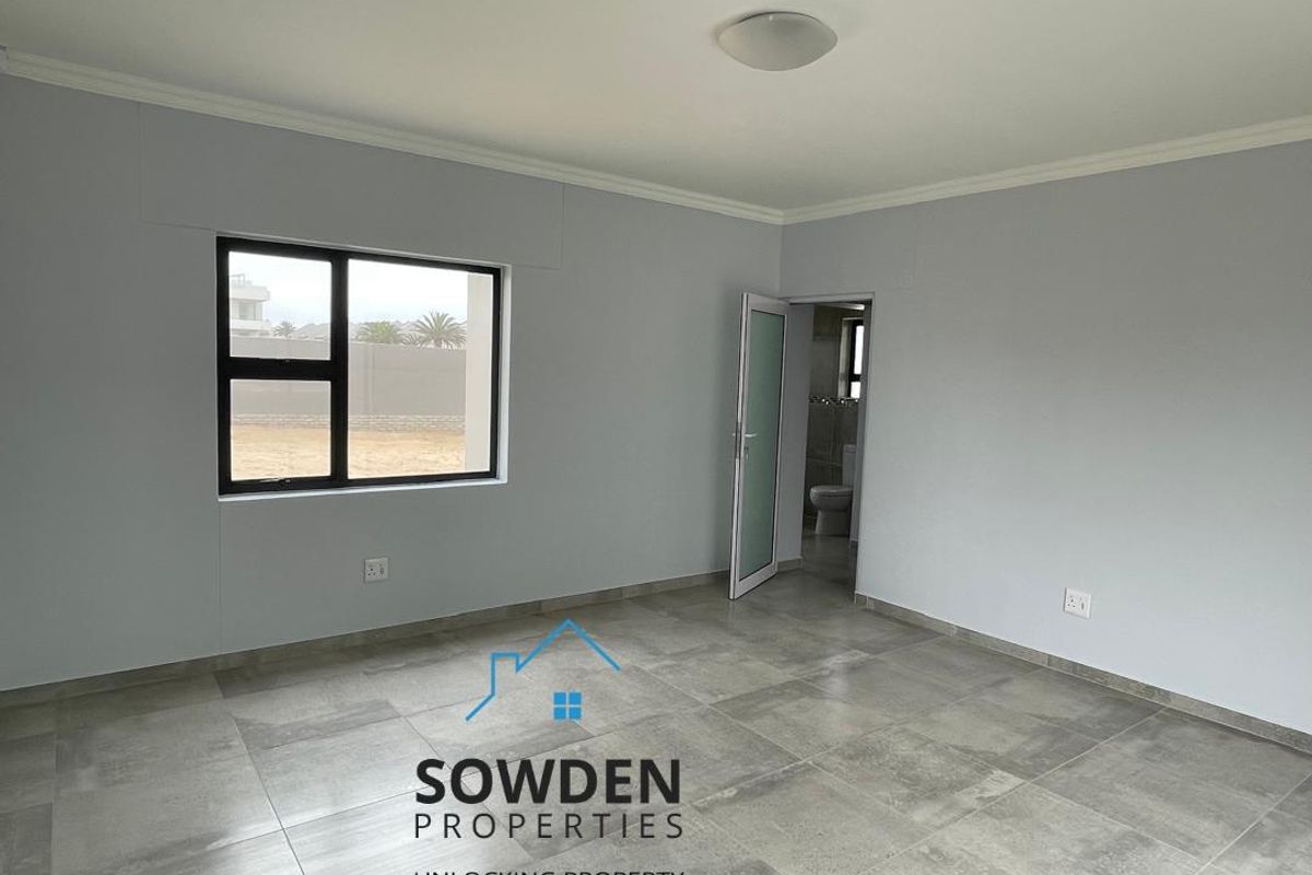 A bright, empty bedroom featuring neutral grey tiled flooring, modern black-framed windows, and an en-suite bathroom access.