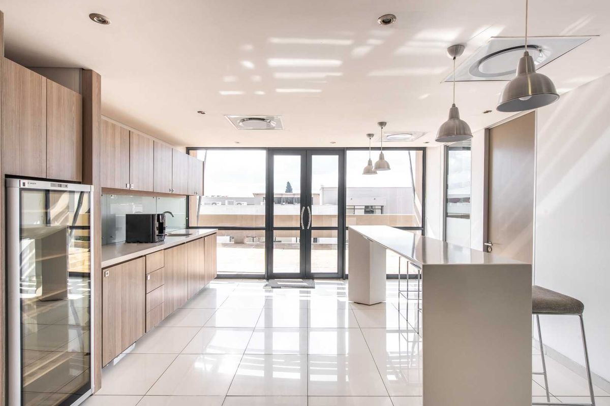 Modern office kitchenette featuring light wood cabinetry, stone countertops, integrated appliances, and a breakfast bar, leading to an outdoor balcony area.