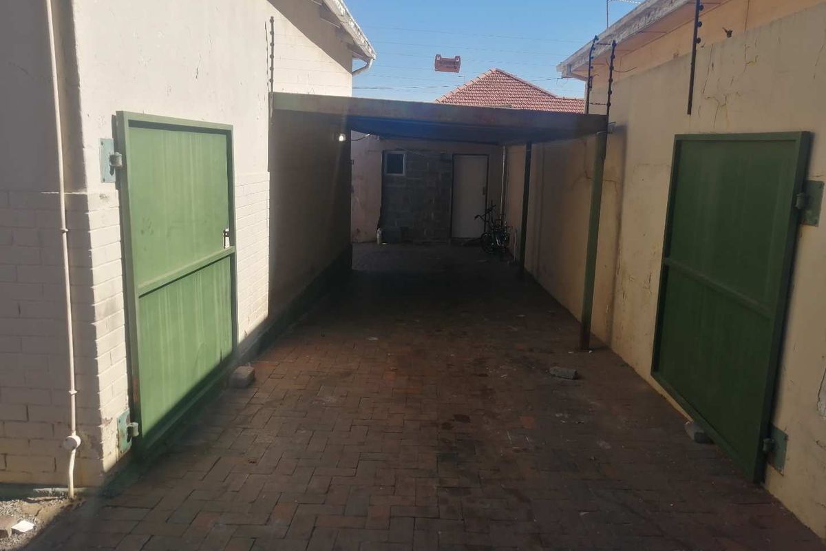 A narrow driveway area featuring a covered carport structure, brick paving, and green metal gates between two building walls.