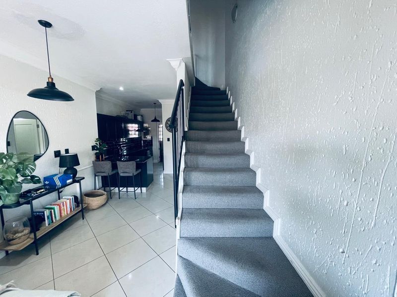 A bright hallway featuring a carpeted staircase with a black metal handrail, leading towards an open-plan kitchen area with tiled flooring.
