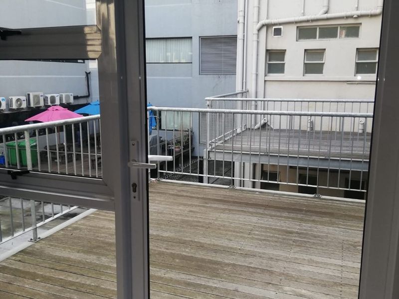A functional outdoor balcony featuring timber decking and a metal balustrade, accessible via an aluminium sliding door, overlooking a neighbouring building courtyard.