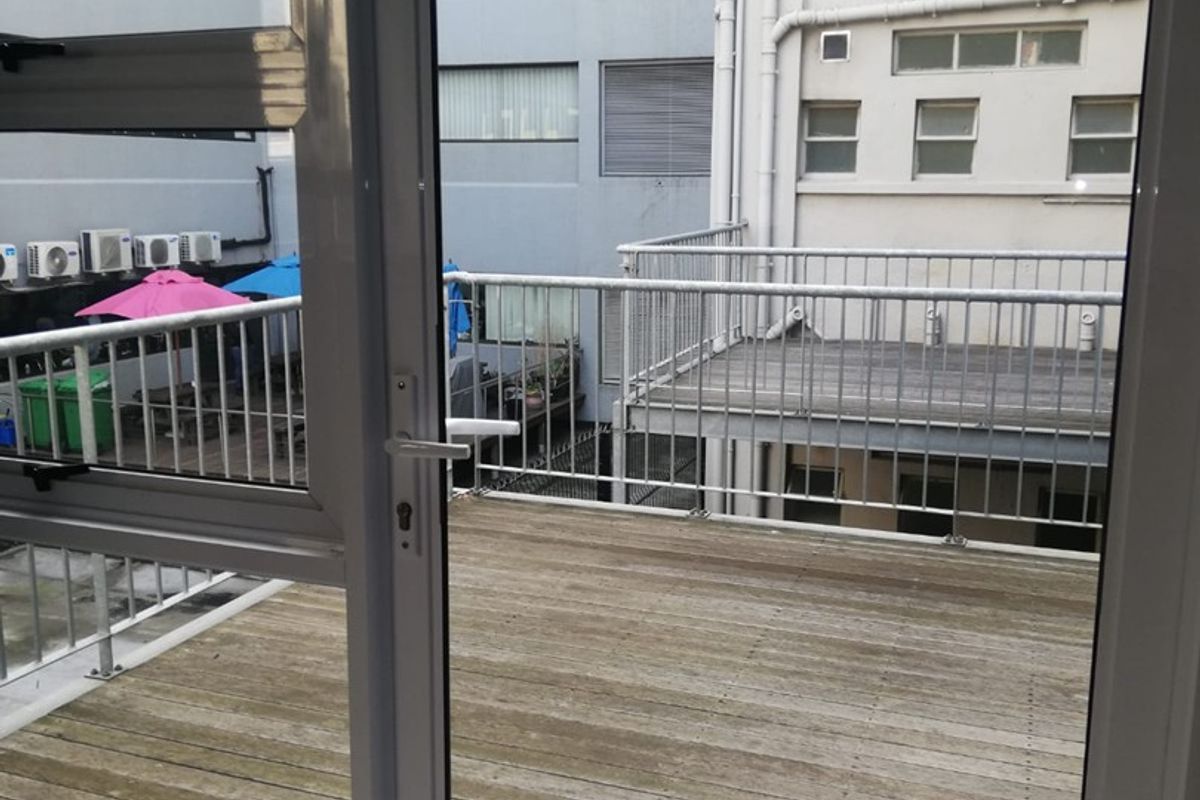 A functional outdoor balcony featuring timber decking and a metal balustrade, accessible via an aluminium sliding door, overlooking a neighbouring building courtyard.