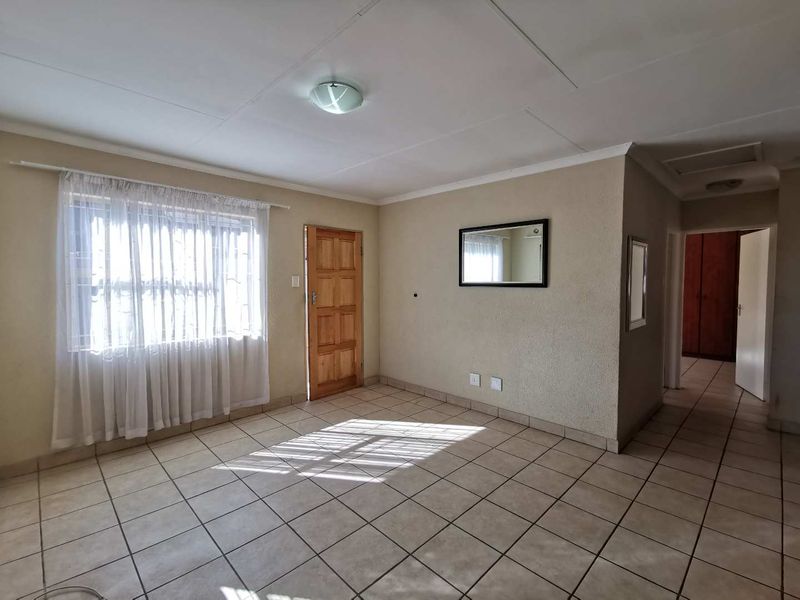 A spacious, empty living area featuring neutral tiled flooring, a timber entrance door, and natural light through a window with security bars.