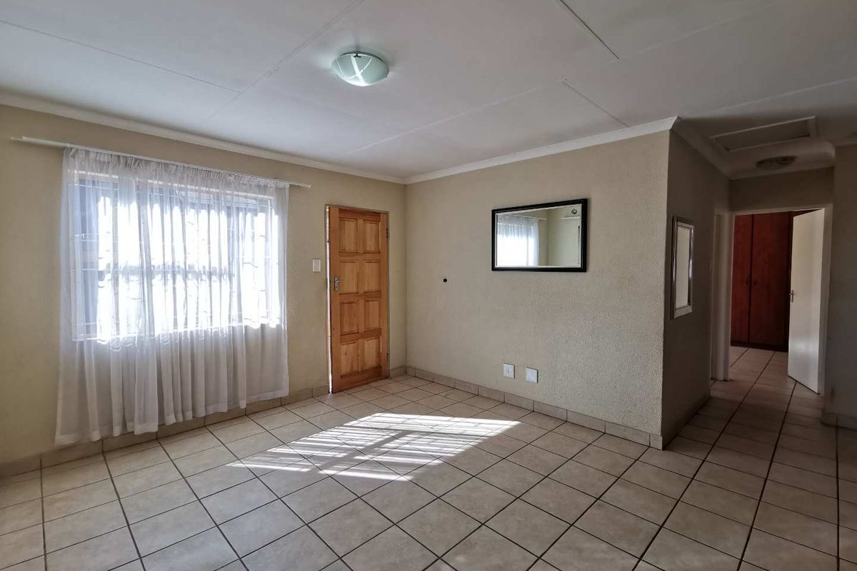 A spacious, empty living area featuring neutral tiled flooring, a timber entrance door, and natural light through a window with security bars.