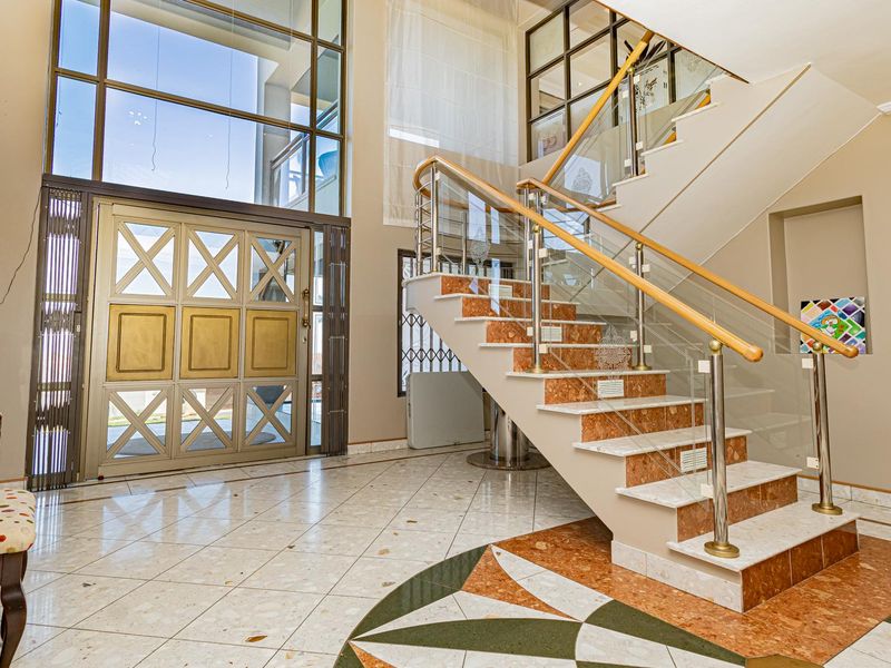 Grand double-volume entrance hall featuring a striking marble staircase with glass balustrades, decorative metal front door, and elegant tiled flooring.